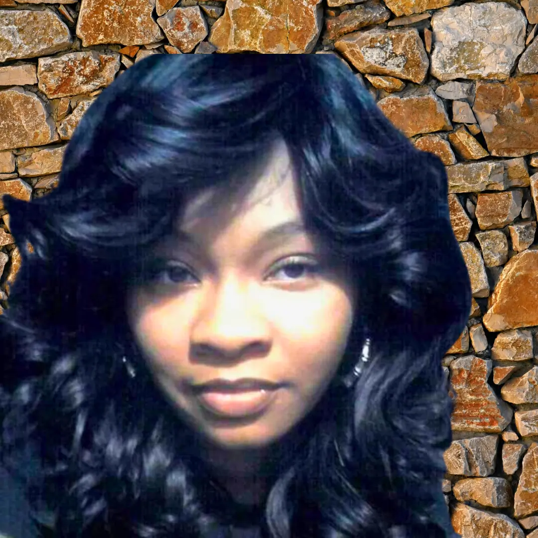 A woman with long, curly black hair poses in front of a stone wall.