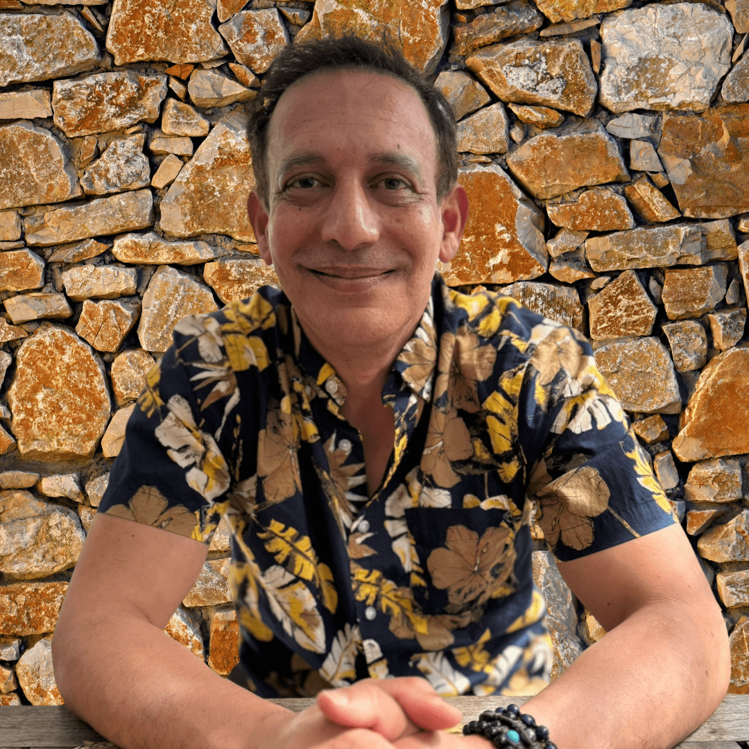 Smiling man in a floral shirt sitting at a table with a stone wall background. Smiling man in a floral shirt sitting at a table with a stone wall background.